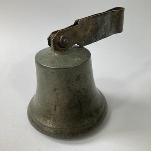 May include: Antique bronze bell with a weathered patina. The bell has a flared base and a hinged handle. The handle is attached with a nut and bolt. The bell is likely made of cast bronze and shows signs of age and use.