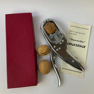May include: A silver-coloured metal Crackerjack nutcracker with a walnut inside, two additional whole walnuts, a rectangular red box, and an instruction pamphlet titled How to get the best from your Three-in-One CRACKERJACK.