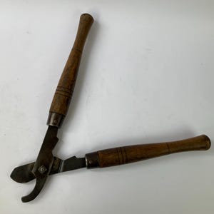 May include: A pair of vintage metal shears featuring two long, cylindrical wooden handles with dark brown wood grain and decorative grooved lines. The metal blades are dark, weathered, and connected by a central pivot bolt mechanism.