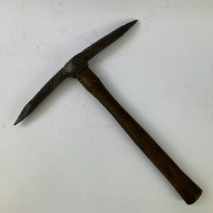 May include: A geological rock hammer featuring a dark, weathered metal head with a pointed pick on one side and a flat chisel end on the other. The tool is attached to a straight, dark brown wooden handle with a smooth, cylindrical finish.