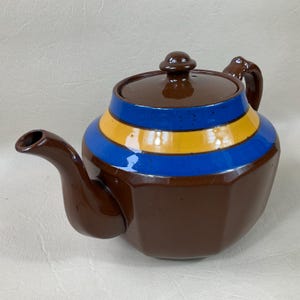 May include: A brown teapot with a blue and yellow striped design. The teapot has a lid and a handle.