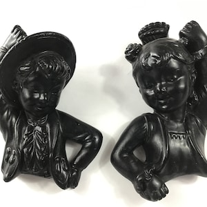 May include: Two black ceramic figurines of a boy and girl in traditional Spanish dress. The boy is wearing a hat and the girl has her hair in a bun. Both figurines are holding their hands up in a dancing pose.