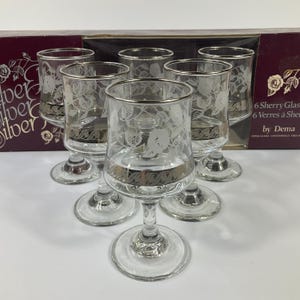 May include: A set of six clear sherry glasses with silver rims and floral etched patterns. The glasses are arranged in a circular pattern, highlighting their elegant shape and detailing. The box behind the glasses reads "6 Sherry Glasses".