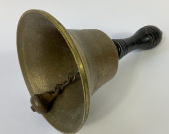 Puede incluir: Una campana de latón con un mango de madera oscura. La campana tiene forma acampanada con un borde dorado y un pequeño badajo colgando en el interior. El mango es de madera oscura barnizada. La campana probablemente se utiliza para señalización o decoración.