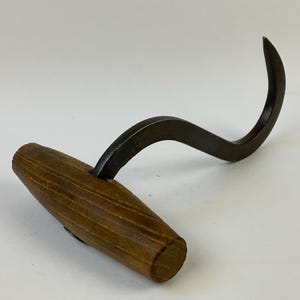 Puede incluir: Un gancho de metal de estilo vintage con una hoja curva de color gris oscuro y un mango de madera. El mango es de color marrón claro con vetas de madera visibles. El gancho está diseñado para colgar o tirar.