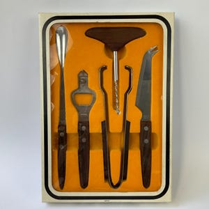 Op de afbeelding: Een barset in een rechthoekige doos met een feloranje binnenkant. De set bevat een lange zilveren lepel, een flesopener, een T-vormige kurkentrekker en een kaasmes, allemaal met donkerbruine houten handvatten met twee zilveren klinknagels.