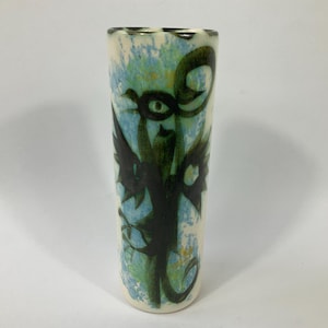 May include: A tall, cylindrical ceramic vase with a cream base and a painted design. The design features a dark green, abstract creature with large eyes and wing-like shapes against a blue and green background. The vase has a slightly flared rim.