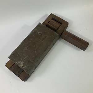 Puede incluir: Un juguete de madera antiguo, también conocido como matraca. El cuerpo rectangular es de madera oscura y una placa de metal. Un mango de madera se extiende desde el lateral. El juguete está diseñado para crear un sonido fuerte y traqueteante al girarlo.
