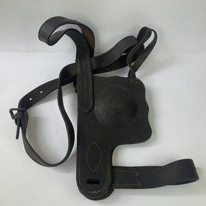 May include: A dark brown leather shoulder holster featuring a molded circular section, adjustable straps with metal buckles, and reinforced stitching along the edges. The harness design includes multiple leather bands intended for securing an object.