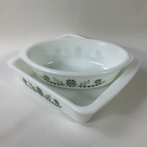 May include: A set of two white ceramic baking dishes with a green floral design. The dishes are nested together, with the smaller dish on top of the larger dish.