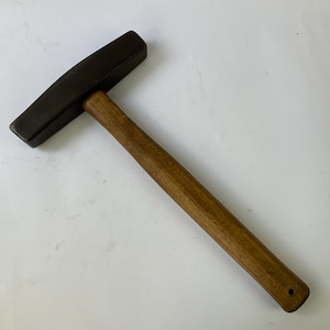 May include: A cross peen hammer featuring a dark gray metal head with a flat, rectangular striking face and a perpendicular wedge-shaped peen. The tool includes a straight, cylindrical wooden handle with a small circular hole at the base.