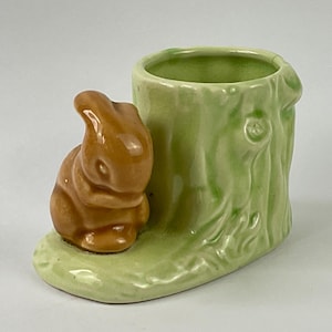May include: A ceramic planter featuring a brown bunny figurine next to a light green tree trunk-shaped container. The planter has a light green base and is likely for small plants or decorative items.