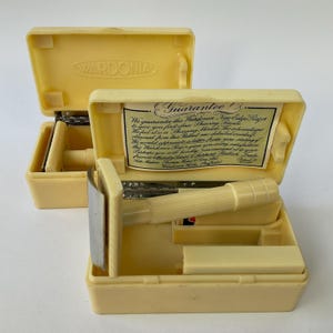 Pode incluir: Duas máquinas de barbear vintage WARDONIA New Edge em estojos abertos de cor creme. Cada estojo contém uma máquina de barbear com cabo creme e lâmina prateada. Um estojo tem um papel com o texto "Guarantee!" e menciona "Shaving Service."