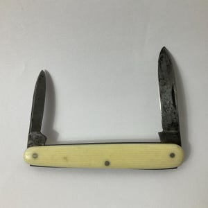 May include: A vintage pocket knife with two blades extended. The blades are a dark, aged metal, and the handle is a cream color with three small rivets. The knife is set against a plain white background.