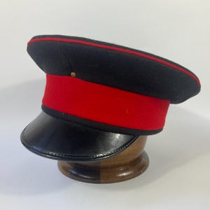 May include: A black and red military-style hat with a shiny black peak. The hat has a red band and trim, and a small gold button on the crown. The hat is displayed on a wooden stand.