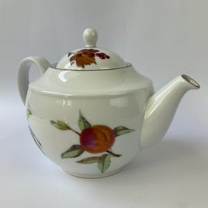 Op de afbeelding: Een witte porseleinen theepot met een handgeschilderd ontwerp van een oranje en rode perzik met groene bladeren op de buik. Het bijpassende deksel is voorzien van een kleine ronde knop en versierd met een trosje rode bessen en bruine bladeren.