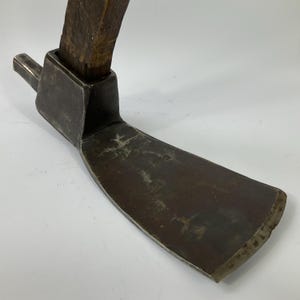 May include: A vintage hoe with a dark, weathered metal blade and a wooden handle. The blade is wide and curved, showing signs of age and use. The handle is attached at a right angle to the blade, suggesting a tool for gardening or farming.