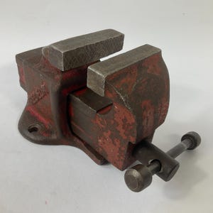 Puede incluir: Un tornillo de banco de metal rojo y gris vintage. El tornillo de banco tiene una base robusta con un orificio para pernos, una mordaza móvil y un mango para apretar. El metal muestra signos de desgaste, lo que le da un aspecto rústico. Ideal para proyectos de carpintería o metalurgia.