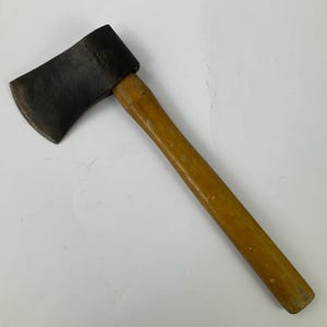 Może przedstawiać: Siekiera ręczna z ciemną, zwietrzałą metalową głowicą z widocznym znakiem stempla oraz gładkim, jasnobrązowym drewnianym trzonkiem. Narzędzie posiada zakrzywioną krawędź tnącą i prosty, cylindryczny trzonek, noszący ślady użytkowania.