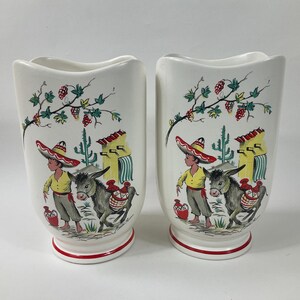 May include: A pair of white ceramic vases with a red rim. Each vase features a colorful illustration of a boy wearing a sombrero and leading a donkey. The scene is set in a desert landscape with a cactus and a small house.