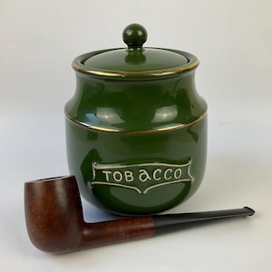 以下が含まれることがあります： 緑色のセラミック製のタバコジャー。蓋と金のトリム付きで、白い文字で「TOBACCO」と書かれています。茶色と黒のタバコパイプがジャーの前に置かれています。
