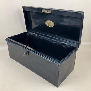 May include: Black metal storage box with an open lid. The box is rectangular with a key lock and a small brass plaque inside the lid. The box is empty and appears vintage. The measurements are in centimetres.