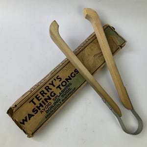 May include: A pair of vintage washing tongs with light-coloured wooden handles and a metal spring mechanism, resting on a rectangular cardboard box labelled Terry's Washing Tongs, Avoid Splashing Boiling Suds, Absolutely Rustproof.