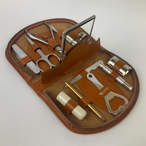 Puede incluir: Neceser de viaje de cuero marrón abierto con varias herramientas de aseo. El kit incluye tijeras, cortaúñas, un espejo, un abrebotellas y otras herramientas metálicas. El neceser tiene cremallera y un interior marrón claro.