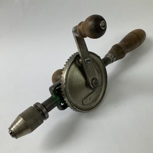 以下が含まれることがあります： メタリックグレーの本体とダークブラウンの木製ハンドルを備えた手動ドリル。この工具はギア機構、前面のローレット加工されたチャック、ギアとチャックの側面に刻印されたFABREXの文字が特徴です。金属製のアームにはMADE IN ENGLANDと400の刻印があります。