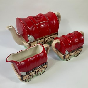 Puede incluir: Un juego de té de cerámica rojo, blanco y marrón con forma de carromatos cubiertos. La tetera, la jarra de crema y el azucarero están decorados con ruedas de carro y una capota de lona roja.