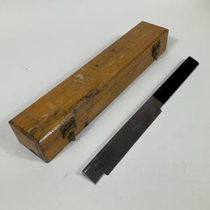 May include: A wooden box with a metal latch and a metal file inside. The box is made of light wood and has a worn finish. The file is made of metal and has a black handle.