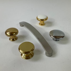May include: Four cabinet knobs in different styles. Two are gold, one is silver, and one is white with a gold rim. The knobs are on a white surface.