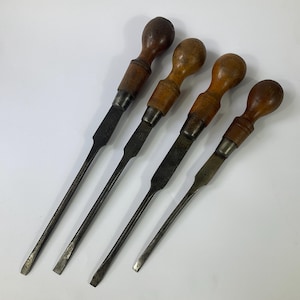 Può includere: Quattro cacciaviti vintage con manici in legno e steli in metallo. I manici sono di diverse tonalità di marrone, di forma arrotondata. Gli steli metallici sono piatti e mostrano segni di usura. Gli utensili sono disposti in diagonale.