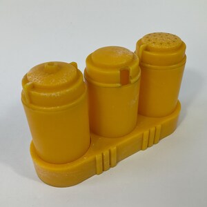 May include: A vintage yellow plastic salt and pepper shaker set. The set includes three cylindrical containers with rounded lids, mounted on a rectangular base. The shaker on the right has a perforated top. The set is made of yellow plastic.
