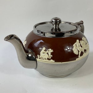 Puede incluir: Una tetera marrón y plateada con un diseño blanco de figuras y una tapa plateada.