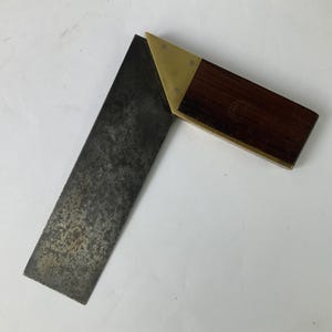 May include: A try square tool featuring a dark, weathered steel blade attached to a wooden handle. The handle is reinforced with brass plates on both sides, secured by three small rivets. The wood has a deep brown tone with a visible grain pattern.