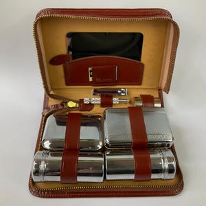 Puede incluir: Neceser de viaje de cuero marrón abierto con cierre de cremallera. El interior presenta un espejo, varias herramientas de aseo y recipientes asegurados con correas de cuero marrón. El neceser está forrado con un material beige claro.