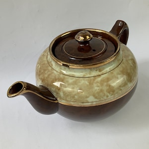 Puede incluir: Una tetera redonda de cerámica con una base de color marrón oscuro y una sección superior de color crema más claro con un patrón moteado similar a las nubes. La tetera incluye una tapa a juego con un pequeño pomo, un pico curvo y bordes dorados.