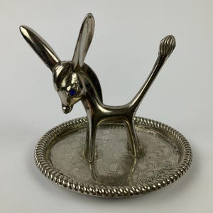 Puede incluir: Figura de burro plateada con ojos azules, de pie sobre una bandeja redonda decorativa. El burro tiene orejas largas y una cola levantada. La bandeja tiene un patrón floral detallado y un borde en forma de cuerda.