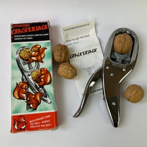 May include: A chrome Crackerjack nutcracker with a walnut inside, accompanied by its original box featuring squirrel illustrations and text, plus an instruction leaflet. The box reads FASCINATING! CRACKERJACK, CRACKS NUTS-CRAB & LOBSTER CLAWS, CRUSHES ICE CUBES, and NUTS STRAIGHT FROM THE SHELL-ALWAYS SWEETEST AND BEST!