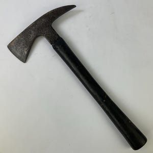 Op de afbeelding: Een vintage brandweerbijl met een donkere, verweerde metalen kop, voorzien van een scherp blad aan de ene kant en een puntige piek aan de andere kant. Het gereedschap is bevestigd aan een lange, rechte, effen zwarte steel voor een goede grip.