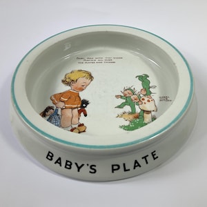 Könnte beinhalten: Eine weiße Baby-Keramikplatte mit hellblauem Rand und dem schwarzen Schriftzug "Baby's Plate". Die Platte zeigt eine farbenfrohe Illustration eines Kindes und Feen. Der Text auf der Platte lautet: "Feen mit winzigen Flügeln, die überall auf den Tellern und Dingen spielen!"