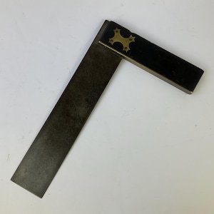 Op de afbeelding: Een metalen winkelhaak met een lang, donker, verweerd stalen blad bevestigd aan een korter, rechthoekig handvat. Het handvat is gemaakt van donker materiaal met een koperen inlegplaat in de vorm van een vierpuntige ster bij de hoek.