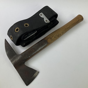 Puede incluir: Una herramienta con una cabeza de metal oscuro que presenta una pica puntiaguda en un lado y una hoja de hacha afilada en el otro, unida a un mango de madera. Junto a ella hay un cinturón de tela negra con ojales metálicos y un clip plateado.