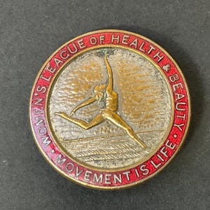 Könnte beinhalten: Eine runde, rot gerandete, goldfarbene Anstecknadel mit einem stilisierten Bild einer tanzenden Frau. Der Text um das Bild lautet "Women's League of Health & Beauty - Movement is Life."