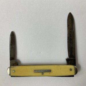 Puede incluir: Una navaja de bolsillo vintage con dos hojas extendidas. La navaja tiene un mango color crema con una virola de metal y una pequeña placa de metal. Las hojas muestran signos de desgaste y envejecimiento, con una apariencia oscura y envejecida.