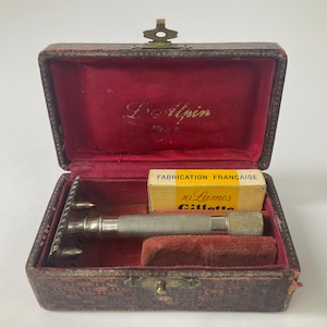 May include: Vintage shaving kit in a leather case. The case is open, revealing a silver razor, a small box of Gillette blades, and a red velvet interior. The case is brown with a brass clasp.