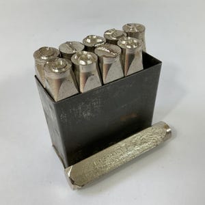 May include: A set of metal letter stamps in a dark metal box. The stamps have various shaped heads and are silver in color. A single stamp is lying outside the box. The text "J.H. SHAND" is visible on the side of the stamp.