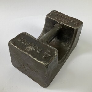 Puede incluir: Peso de hierro fundido gris oscuro, marcado "4 POUNDS" en un extremo y "J. RHODES & SONS" en el otro. El peso tiene un asa. El objeto probablemente se utiliza para pesar. Aproximadamente 1,8 kg.
