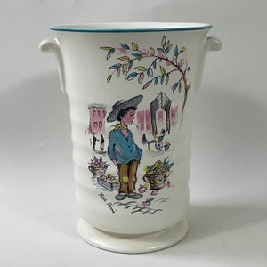 Può includere: Vaso in ceramica bianca con due manici, con un'illustrazione colorata di un ragazzo che vende frutta in una scena di strada. Il vaso ha un bordo blu e verde intorno alla parte superiore e le parole "Petit Fleur" nella parte inferiore.